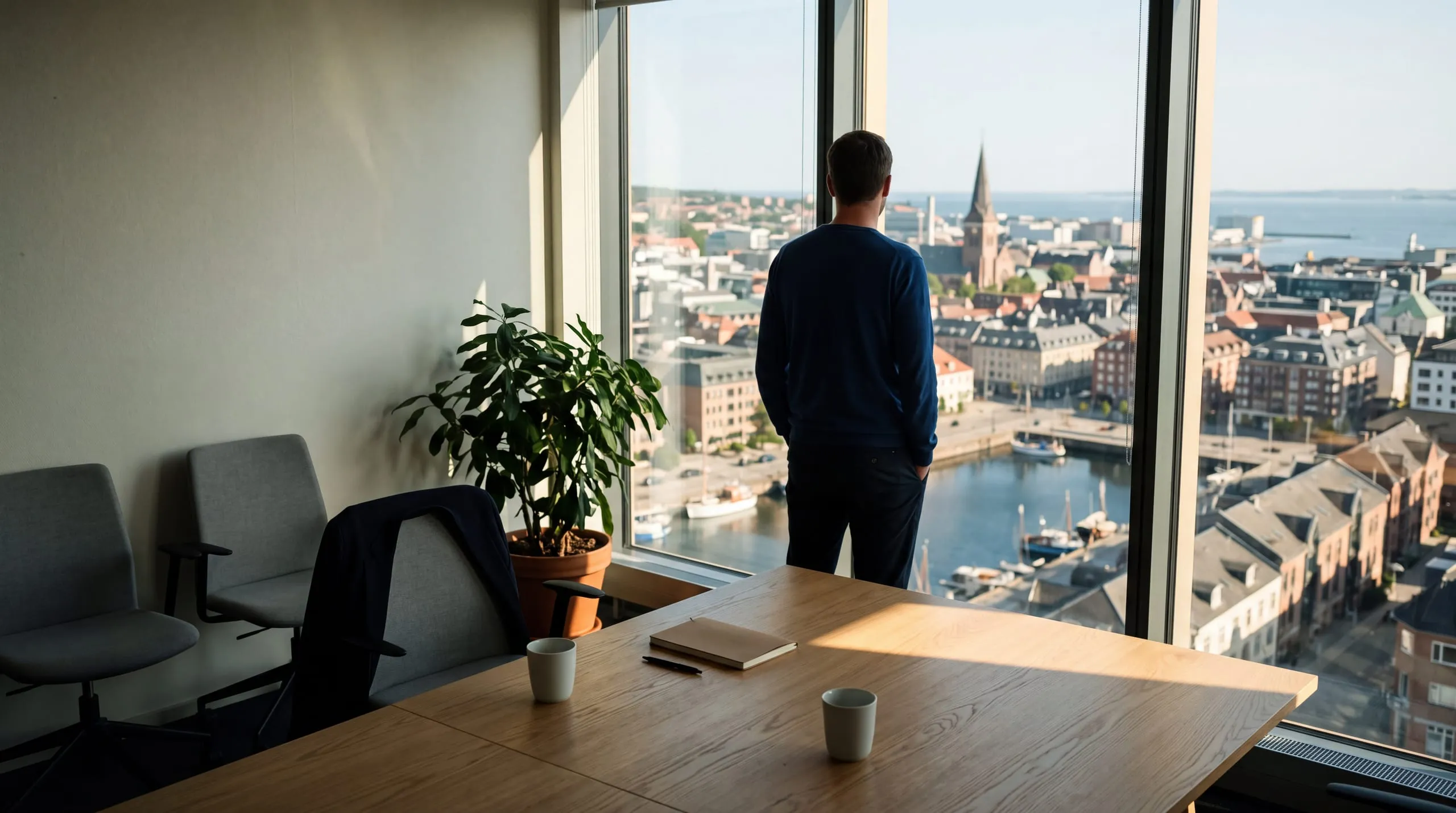 A meeting room with a window wall facing a city and water, morning light on an empty table, a person seen from behind at the window.